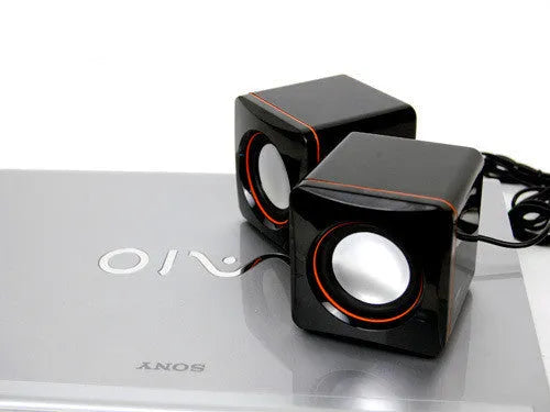 Small black square USB-powered 2.0 computer speaker on a desk next to a laptop