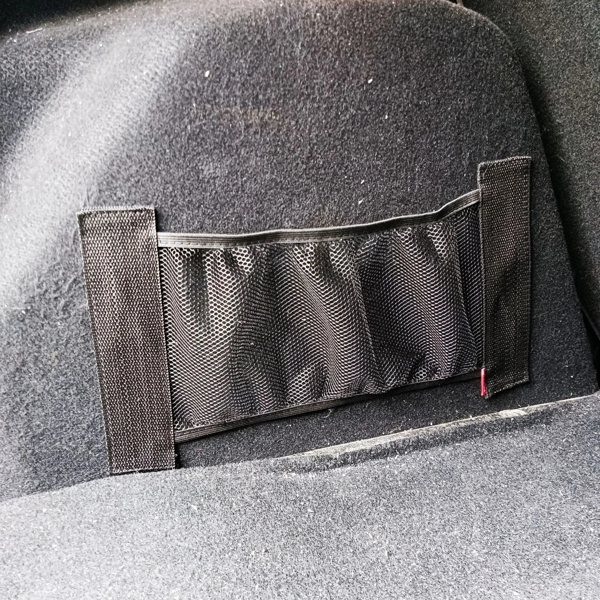 Black elastic polyester storage net stretched in a car boot, holding a water bottle and cloth.