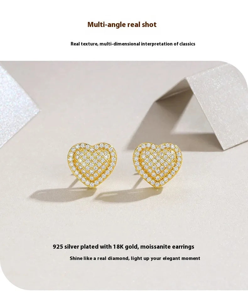 A pair of gold-coloured heart-shaped moissanite stud earrings on a plain background.