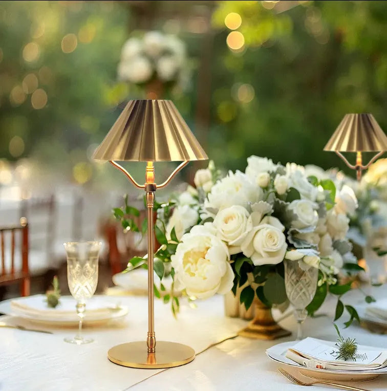Gold alloy desk lamp with a cylindrical shade, shown lit with warm white light on a wooden desk.