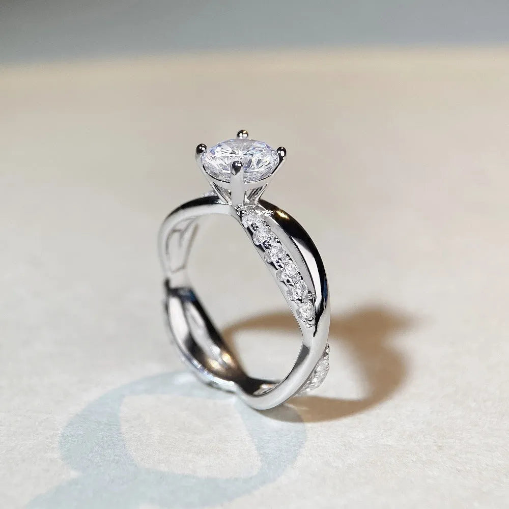 Women's silver ring with a 1 karat moissanite stone in a four-claw twisted arm setting, showing a geometric cross design.