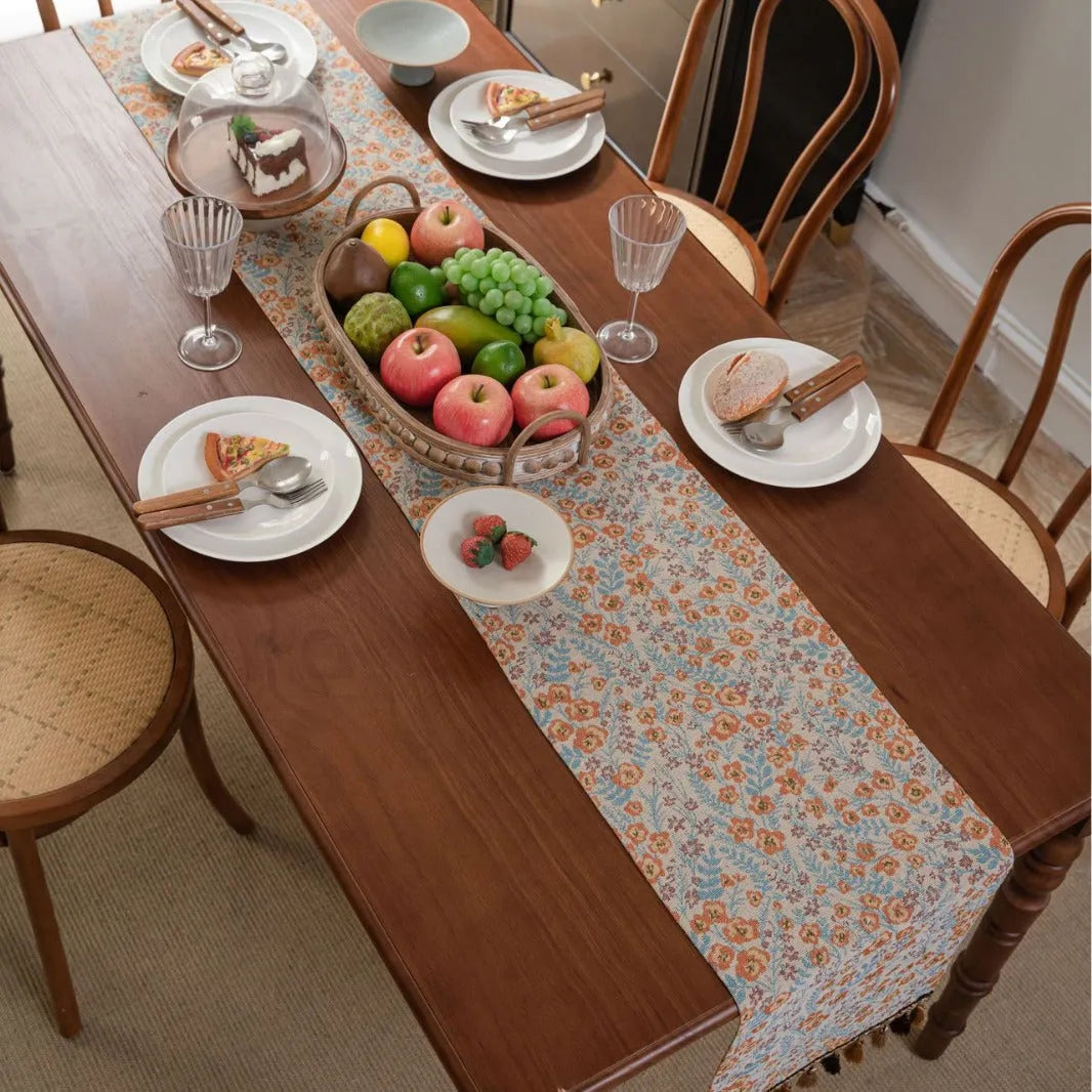 Beige Jacquard table runner with long fabric tassels and floral leaf pattern laid on a wooden table.
