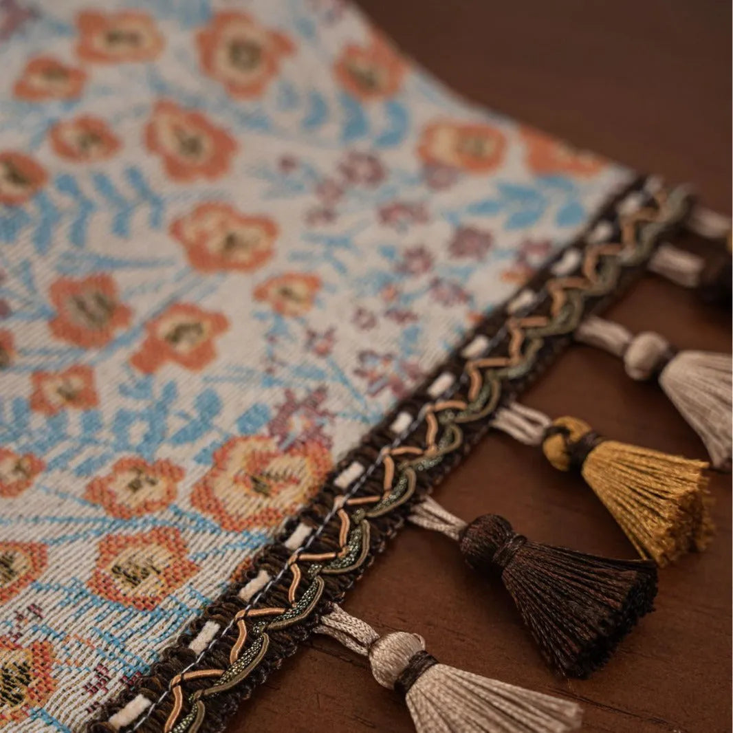 Beige Jacquard table runner with long fabric tassels and floral leaf pattern laid on a wooden table.