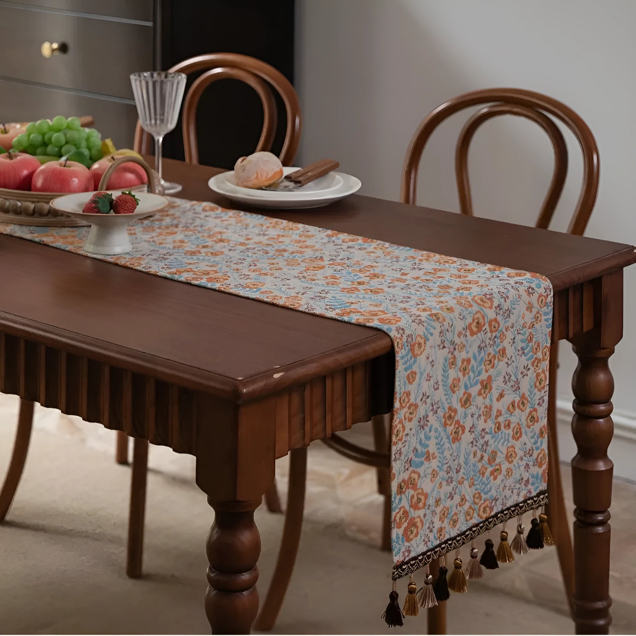 Beige Jacquard table runner with long fabric tassels and floral leaf pattern laid on a wooden table.