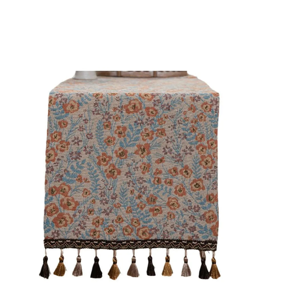 Beige Jacquard table runner with long fabric tassels and floral leaf pattern laid on a wooden table.