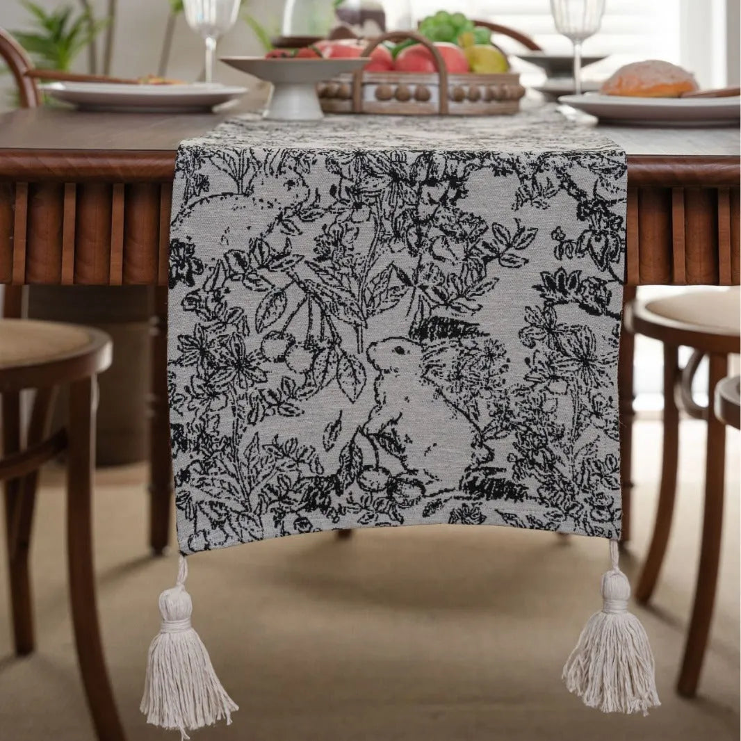 Beige Jacquard table runner with long fabric tassels and floral leaf pattern laid on a wooden table.