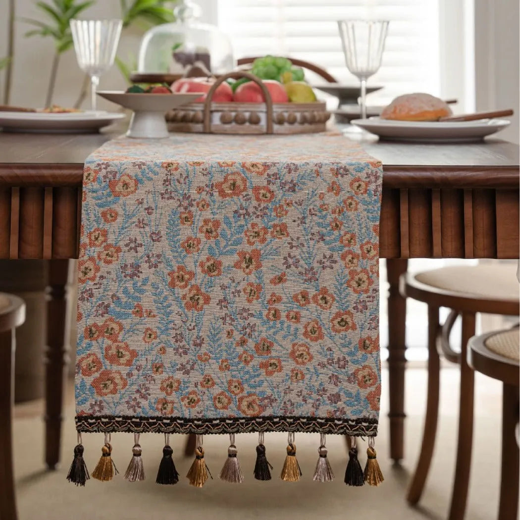 Beige Jacquard table runner with long fabric tassels and floral leaf pattern laid on a wooden table.