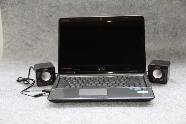 Small black square USB-powered 2.0 computer speaker on a desk next to a laptop