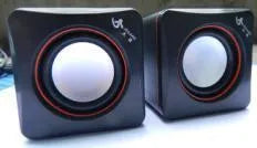 Small black square USB-powered 2.0 computer speaker on a desk next to a laptop